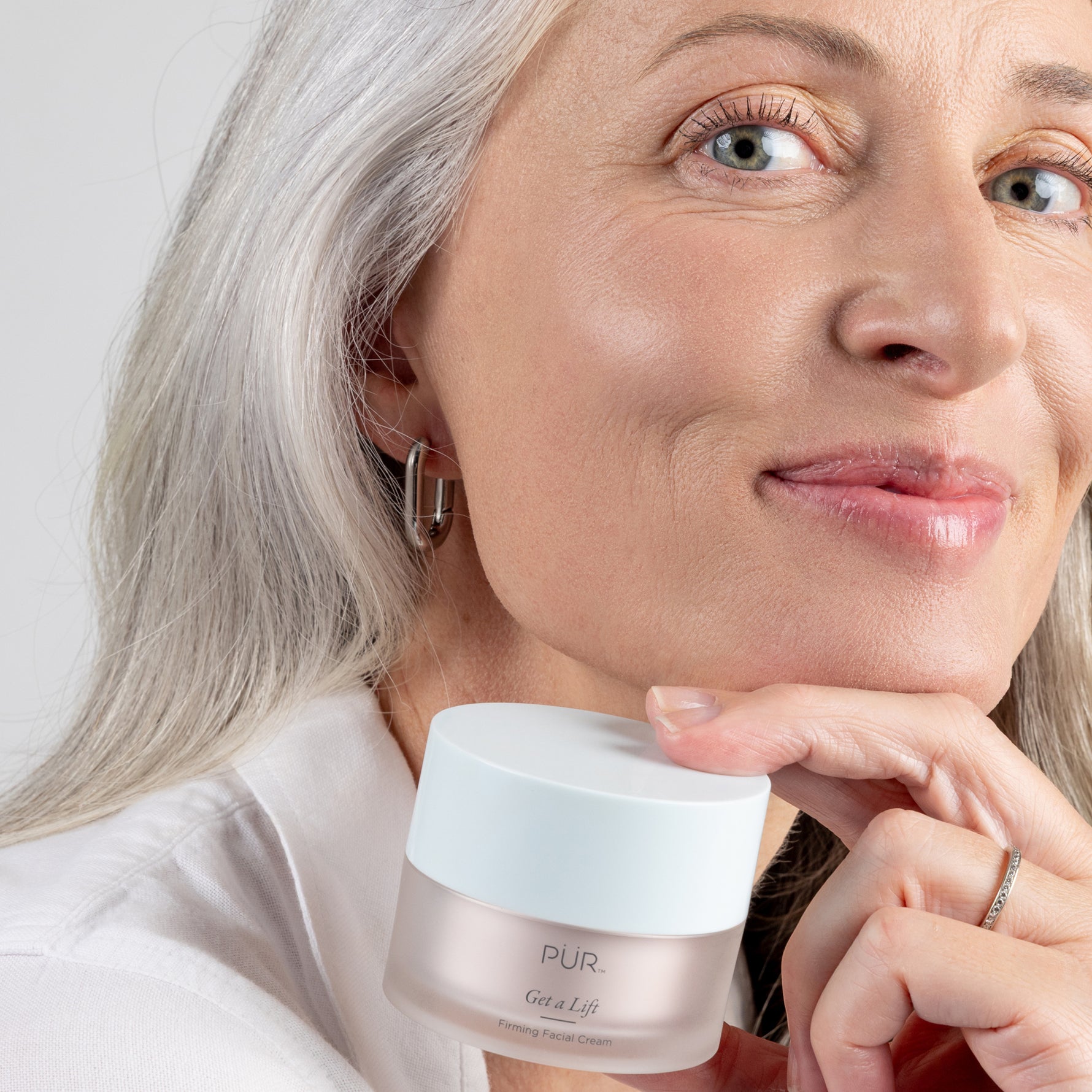 Smiling mature woman holding PÜR Get A Lift Firming Facial Cream, showcasing firm, radiant-looking skin