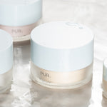 PÜR Tropical C Moisturizer jar with other skincare jars on a soft reflective surface, highlighting clean, minimal packaging and glossy white lids.